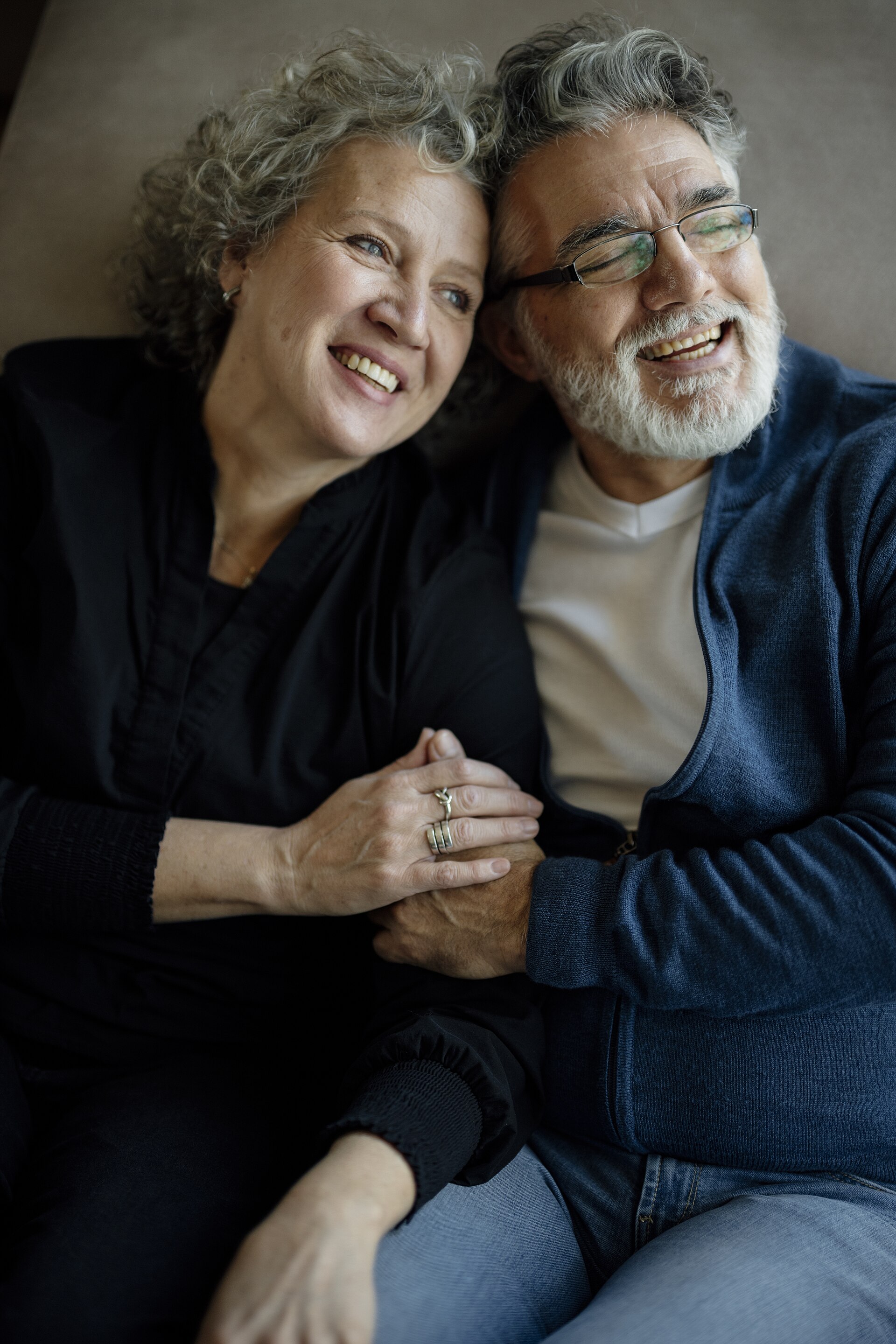 A smiling older couple laughing together on a couch, holding hands